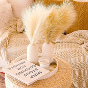 Decorative setup with pampas grass, books, and a candle on a woven stool.