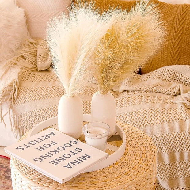 Decorative setup with pampas grass, books, and a candle on a woven stool.