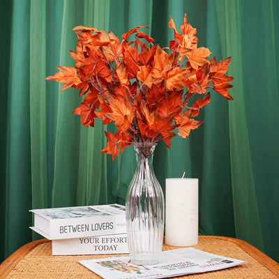Bouquet of orange flowers in a clear vase on a table with books and a candle against a green curtain background.