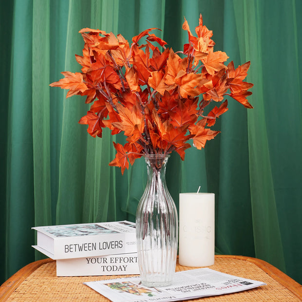 Bouquet of orange flowers in a clear vase on a table with books and a candle against a green curtain background.