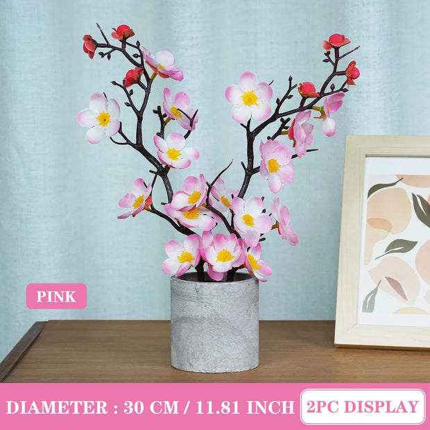 Pink artificial flower branch in a pot on a wooden surface with a light blue wall and framed picture in the background.