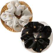 Set of two scrunchies, one white with gold stars and one black with gold stars, on a woven and plain background.