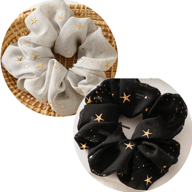 Set of two scrunchies, one white with gold stars and one black with gold stars, on a woven and plain background.