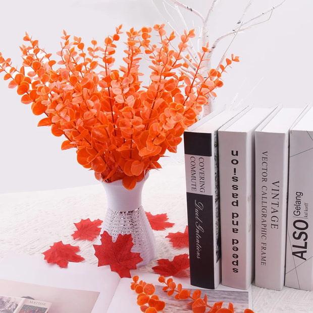 Orange artificial leaves in a white vase with books and red leaves on a white surface