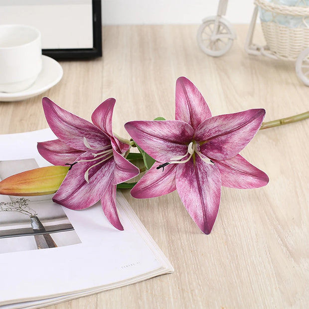 Two pink lilies on a wooden surface with a magazine and decorative items in the background.