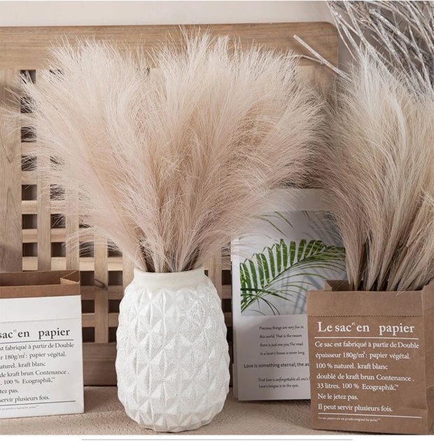 Decorative arrangement with pampas grass in a white vase and brown paper bag on a wooden surface.