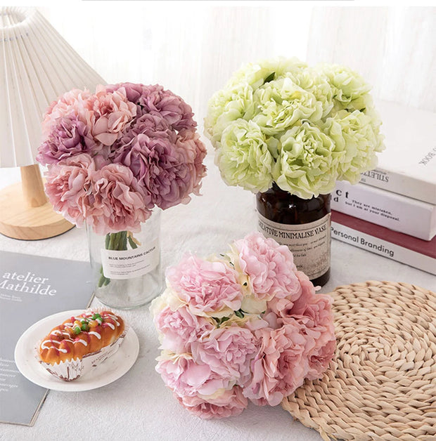 Three flower arrangements in glass vases on a table with books and a pastry.