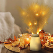 Decorative setup with a jar lantern, dried grasses, and candles on a wooden surface.