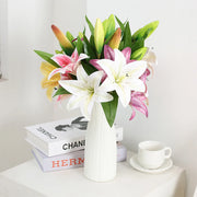 Bouquet of flowers in a white vase on a table with books and a cup in the background.