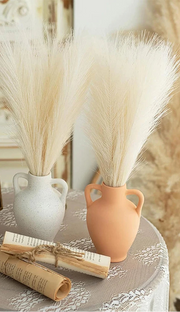 Decorative setting with vases of dried grasses, a framed portrait, and a rolled-up paper on a textured surface.