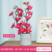 Artificial flower branch with pink flowers in a pot on a table, with a framed picture in the background.