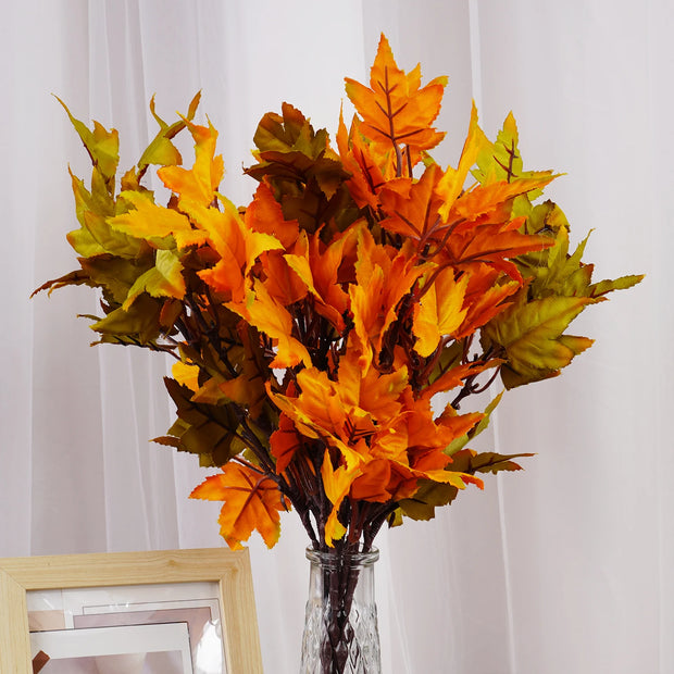 Bouquet of orange and green artificial leaves in a clear glass vase against a white curtain background.