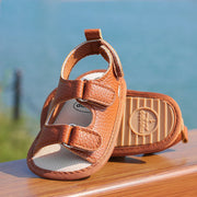 Brown baby sandals with a visible brand logo on a wooden surface with a blurred natural background.