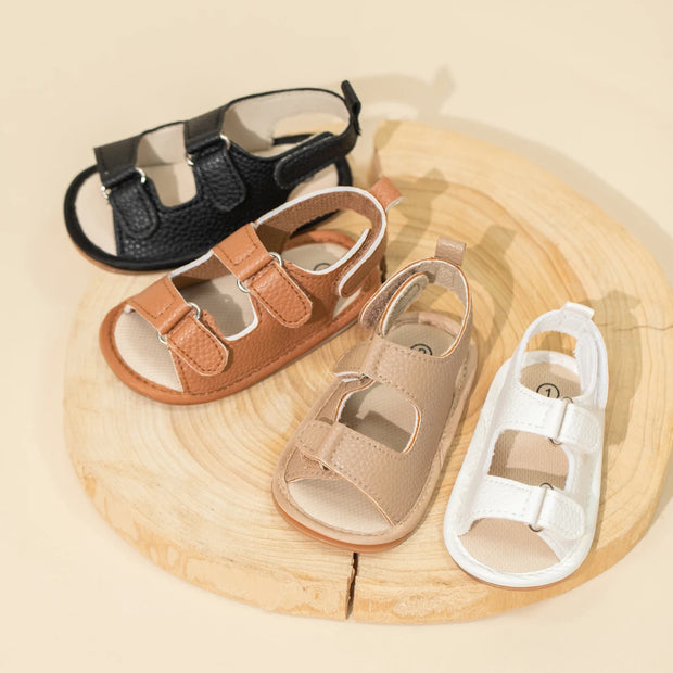Set of four children's sandals in black, brown, beige, and white on a wooden surface.
