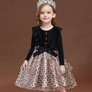 Young girl wearing a black cardigan over a polka dot dress with a tiara on a brown background