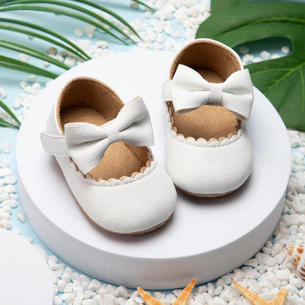 White baby shoes with bows on a white platform surrounded by pebbles and green leaves.