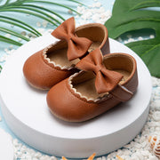 Brown leather baby shoes with bows on a white platform with green leaves in the background