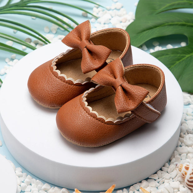 Brown leather baby shoes with bows on a white platform with green leaves in the background