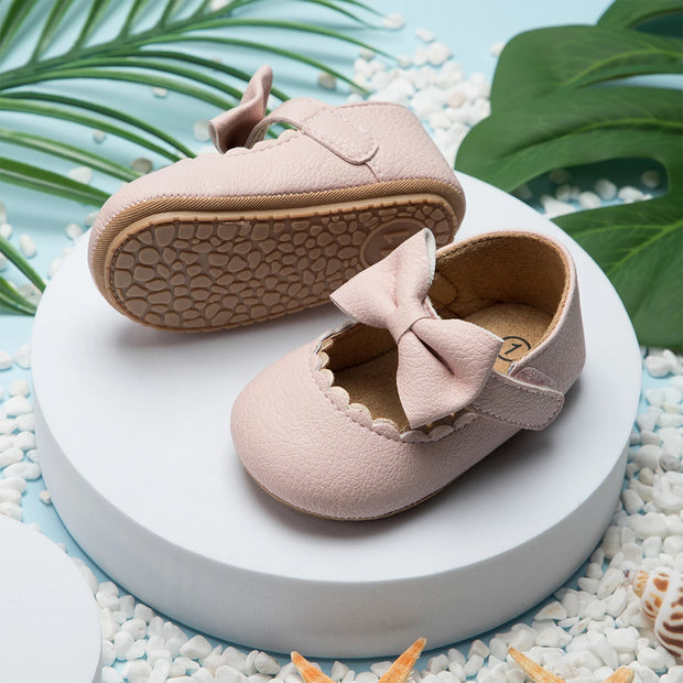 Pair of pink baby shoes with a bow on a white platform with green leaves in the background