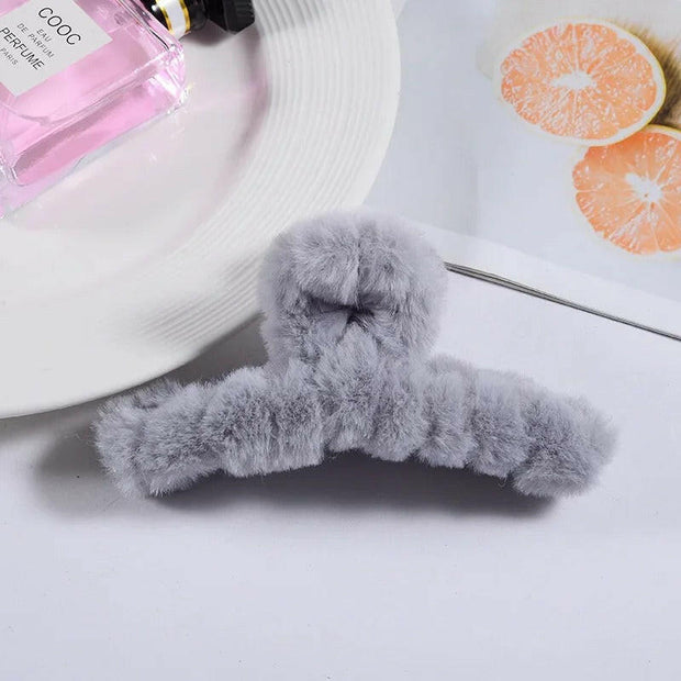 Gray fluffy hanger on a white surface with a bottle of pink perfume and a white hat in the background.