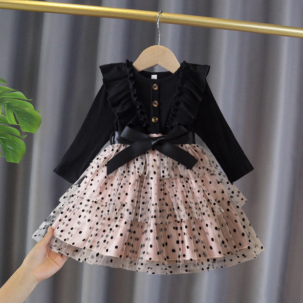 Children's outfit with black top and polka dot skirt on a hanger against a gray curtain background.