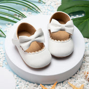 White baby shoes with bows on a white platform surrounded by pebbles and green leaves.