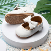 White baby shoes with a bow on a white platform with green leaves in the background