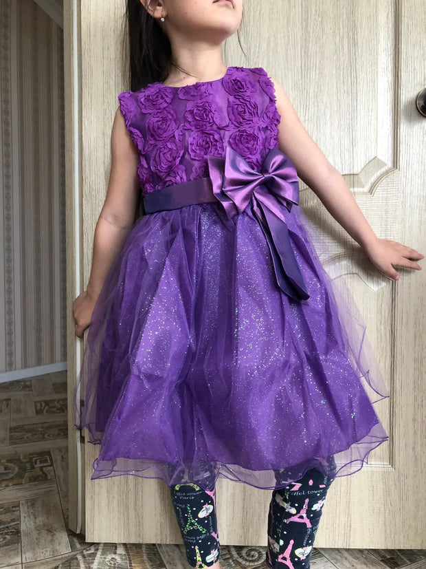 Child wearing a purple dress with floral details and a large bow, standing in front of a wooden door.