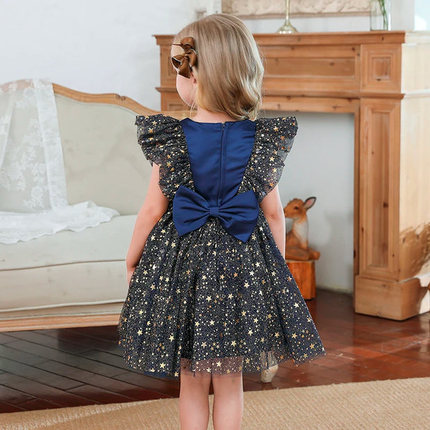 Young girl in a sparkly dress with a blue bow back, standing in a room with a wooden bench and fireplace.