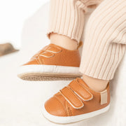 Brown children's shoes with velcro straps worn by a child in light-colored pants.