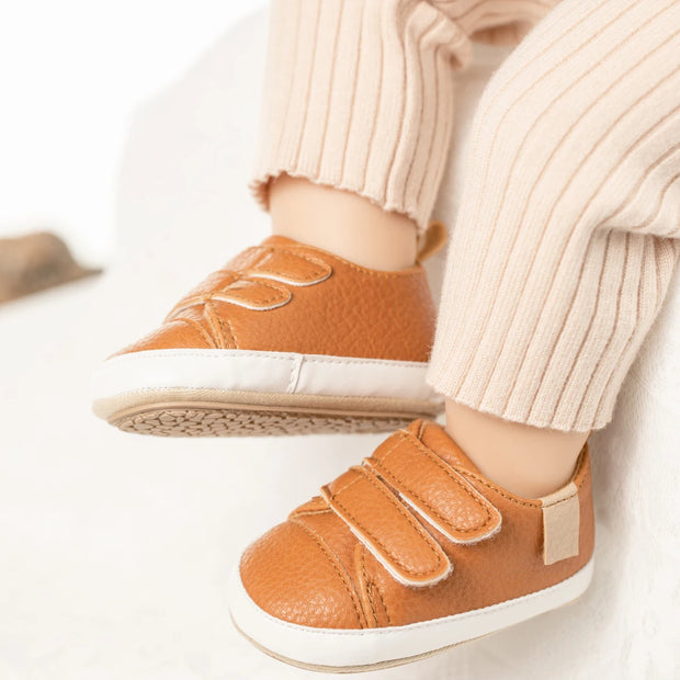 Brown children's shoes with velcro straps worn by a child in light-colored pants.
