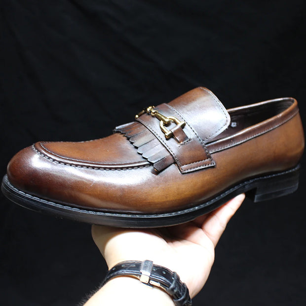 Brown leather loafer with a gold buckle held by a hand against a black background