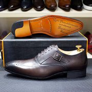 Brown leather dress shoe with gold buckle on a textured surface, brand name visible