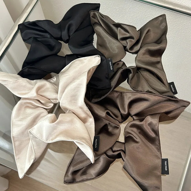 Set of four satin scrunchies in black, beige, and brown on a reflective surface.