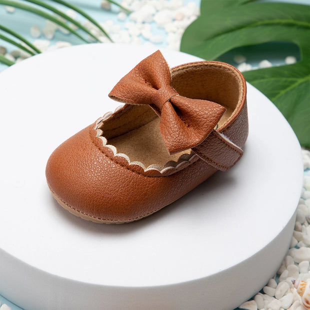 Brown leather baby shoe with a bow on a white surface with green leaves in the background