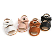 Four pairs of baby sandals in white, pink, brown, and black with bow details on a white background.