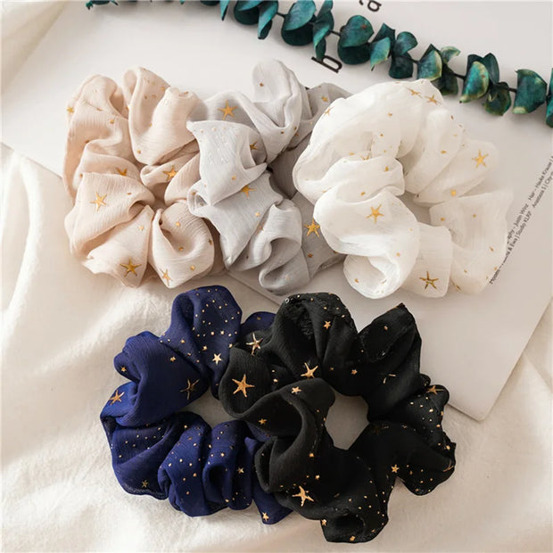 Set of four scrunchies in beige, gray, white, blue, and black with star patterns on a light background.