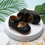 Pair of black leather baby shoes with bows on a white platform with decorative elements.