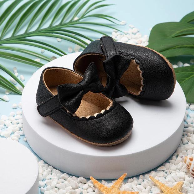 Pair of black leather baby shoes with bows on a white platform with decorative elements.