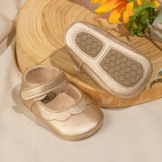 Gold baby shoes with a strap on a wooden surface with flowers.