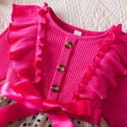Pink ribbed cardigan with ruffled details and a large bow on a textured surface