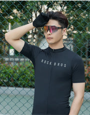 Person wearing a black cycling jersey with sunglasses, standing in front of a chain-link fence.