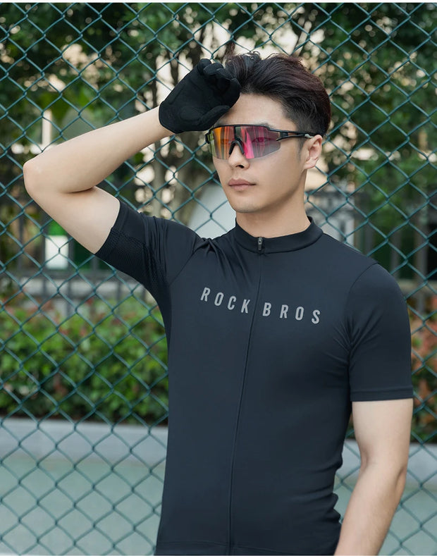 Person wearing a black cycling jersey with sunglasses, standing in front of a chain-link fence.