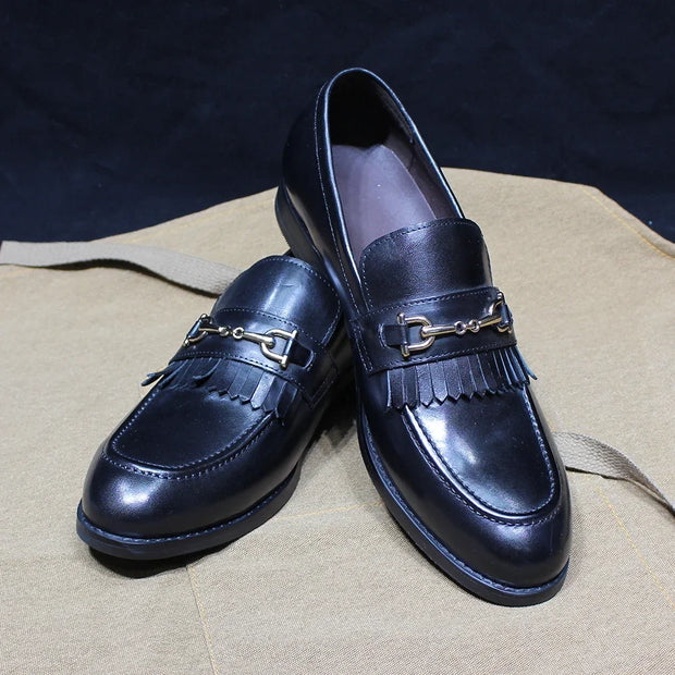 Pair of black loafers with fringe and gold accents on a beige surface.