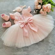 Pink baby dress with a large bow, matching shoes, and headband on a gray surface with flowers.