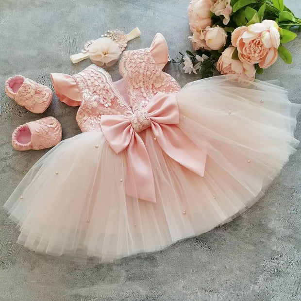 Pink baby dress with a large bow, matching shoes, and headband on a gray surface with flowers.