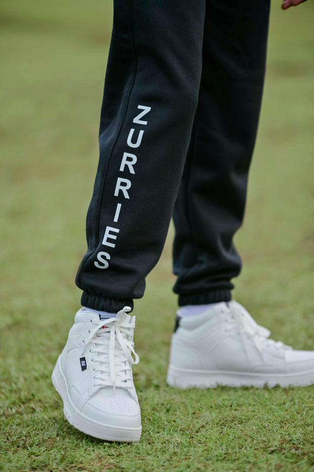 Black jogger pants with 'ZURRIES' text worn by a person on grass.